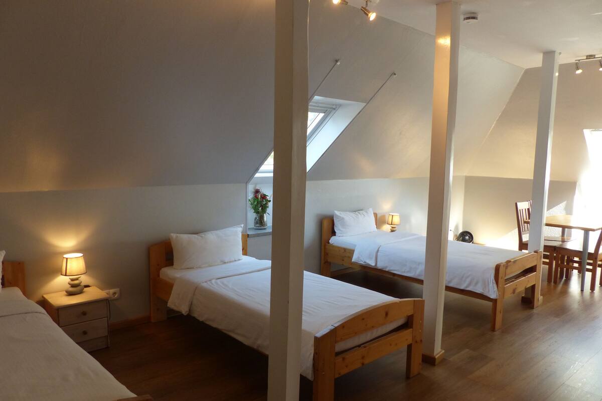 Ein Zimmer mit drei Holzbetten, einem Holztisch und einem Stuhl. Ein Fenster bietet Blick ins Freie.