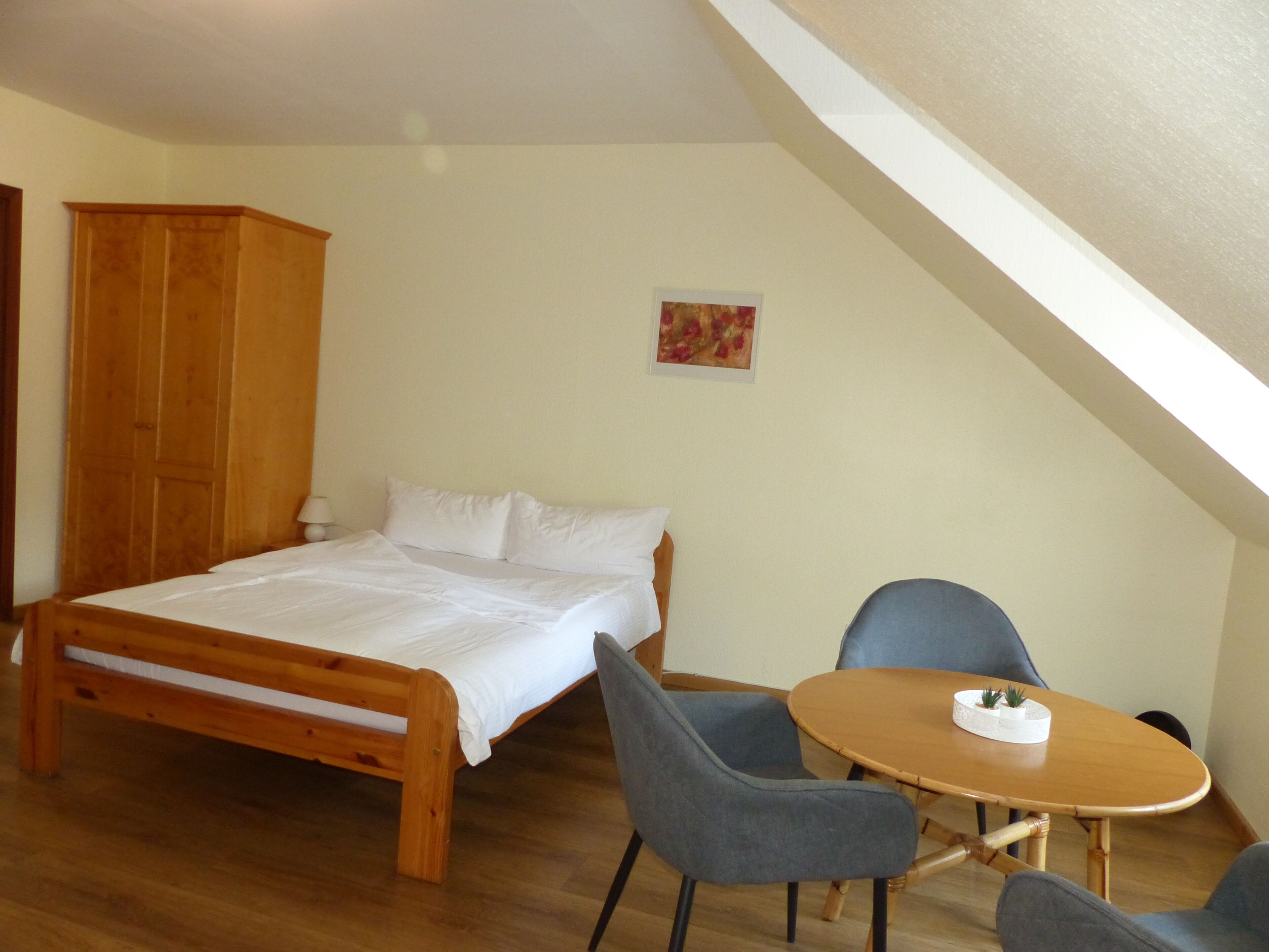 Une chambre avec un lit en bois, deux chaises grises, une petite table ronde ornée d’une plante et une armoire en bois.