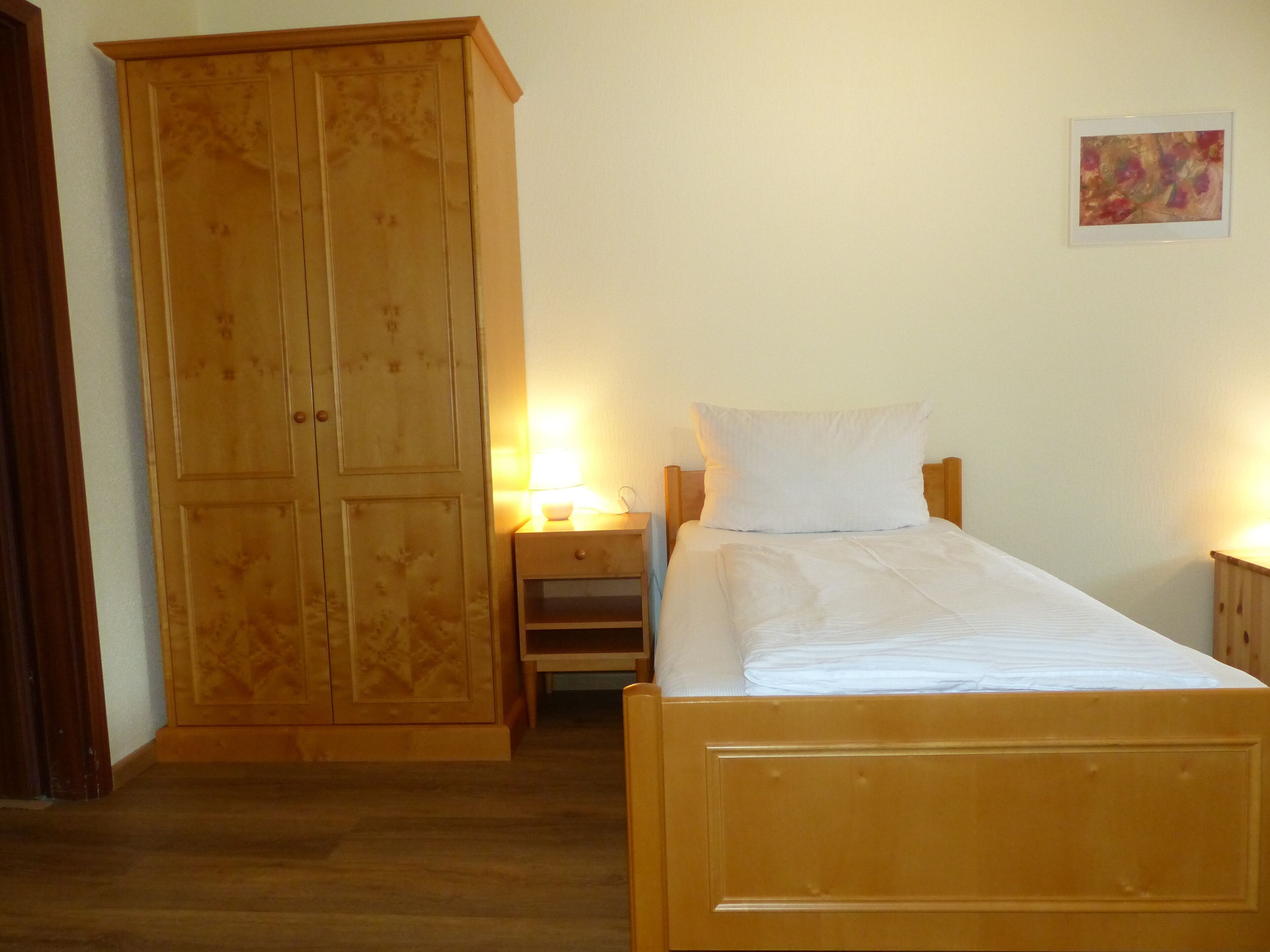 Una habitación con una cama de madera, una mesita de noche con lámpara, un armario y un cuadro en la pared.