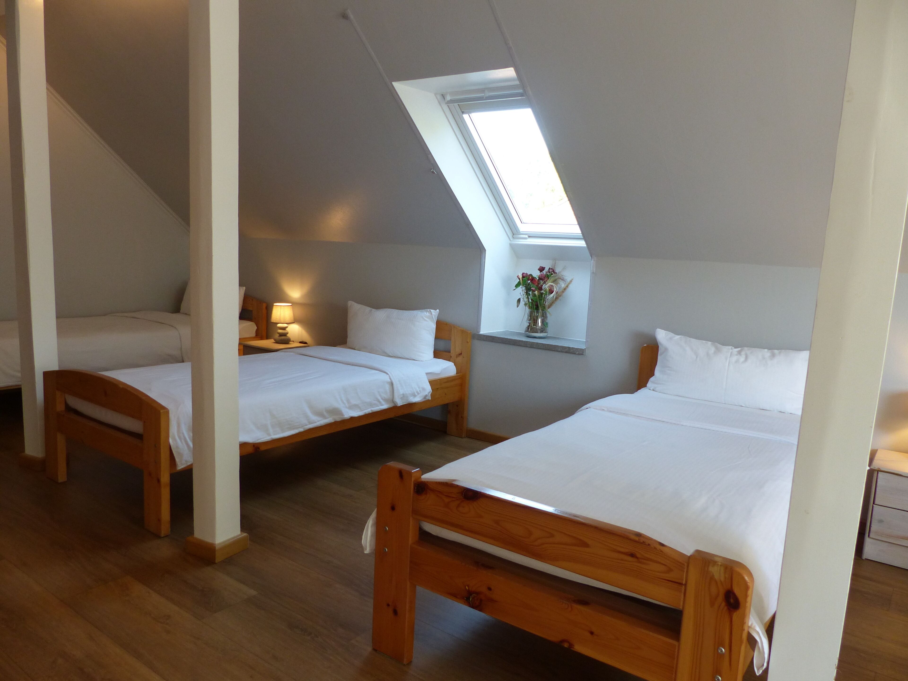 A room with two wooden beds, a skylight, a bedside table with a lamp, and a small window with a vase of flowers.