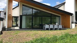 A modern house with a large glass sliding door, a patio with white chairs, and a wooden pergola. - Design Villa Winterberg: Luxury vacation home directly on the slopes/meadow (Winterberg)
