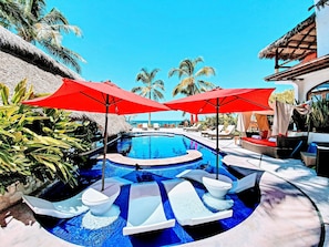 A pool area with lounge chairs and umbrellas, surrounded by palm trees and a clear sky.
