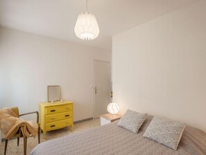 A bedroom with a bed, a yellow dresser, a chair, a bedside table with a lamp, and a door. - L'Amiral by Interhome (Cagnes-sur-Mer)