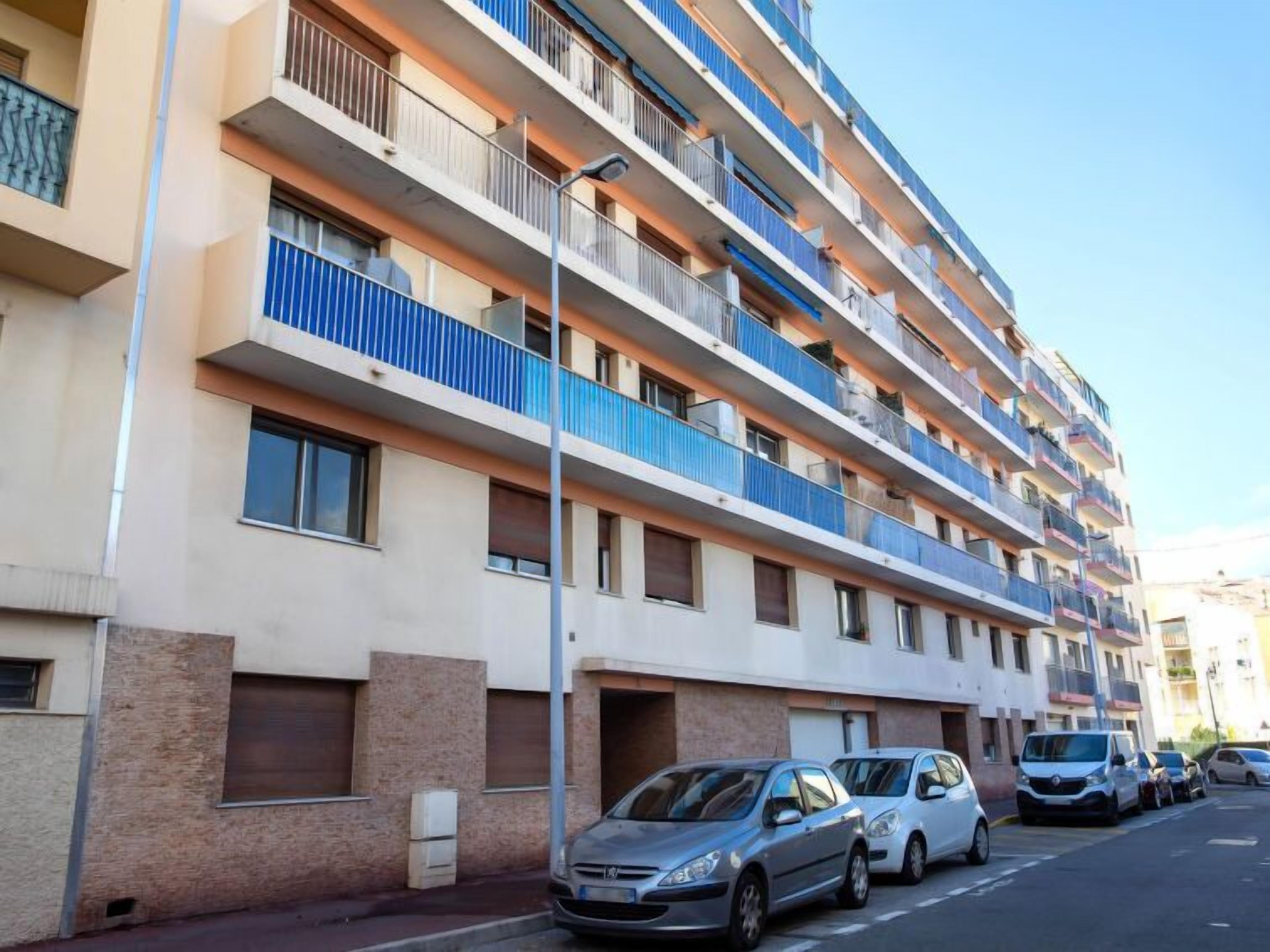 A multi-story residential building with balconies, a parking area, and a clear sky.