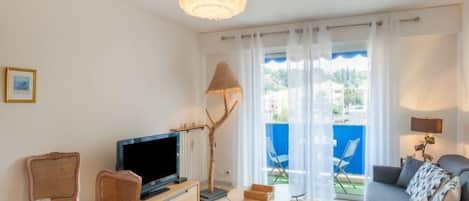 A modern living room with a flat-screen TV, a sofa, a dining table with chairs, and a balcony with a view of the sea.