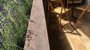 Una terraza de madera con un largo banco, rodeada de hierba verde alta y flores moradas.