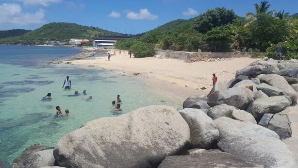 A beach with clear turquoise water, large rocks in the foreground, and people swimming and walking on the shore. - Studio Bougainvillier Style Bohème (Marigot)