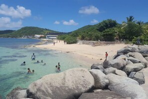 Une plage avec une eau turquoise et limpide, de gros rochers au premier plan, et des gens qui se baignent et se promènent sur le rivage.