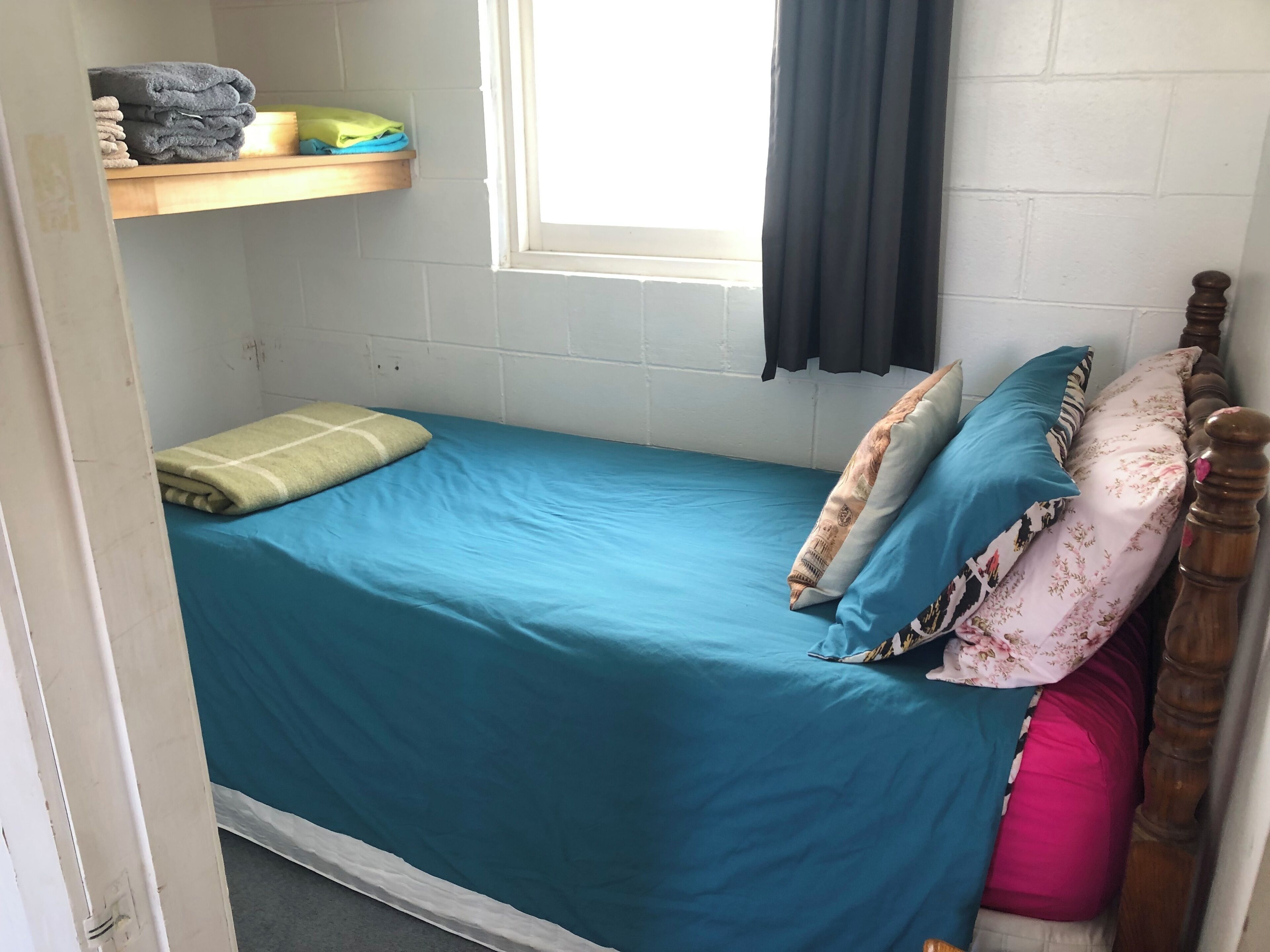 A small, single-bed room with a blue bedspread, a wooden headboard, and a window with a black curtain.