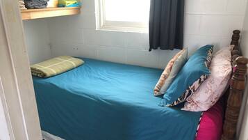 A small, single-bed room with a blue bedspread, a wooden headboard, and a window with a black curtain.