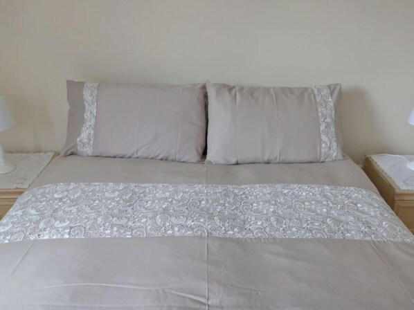 Un lit avec une couvre-lit beige et deux oreillers, une table de chevet avec une lampe et une tête de lit en bois.