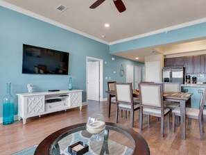A modern living room with a TV, white entertainment unit, and a dining area with wooden chairs and a glass coffee table.