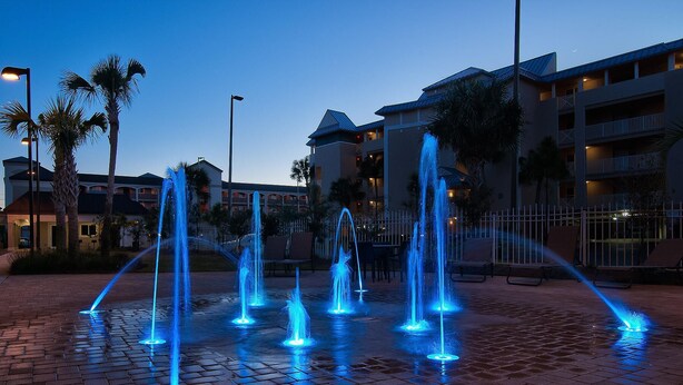 Escena nocturna con varias fuentes iluminadas con luces azules, rodeadas de palmeras y una zona pavimentada con ladrillos.