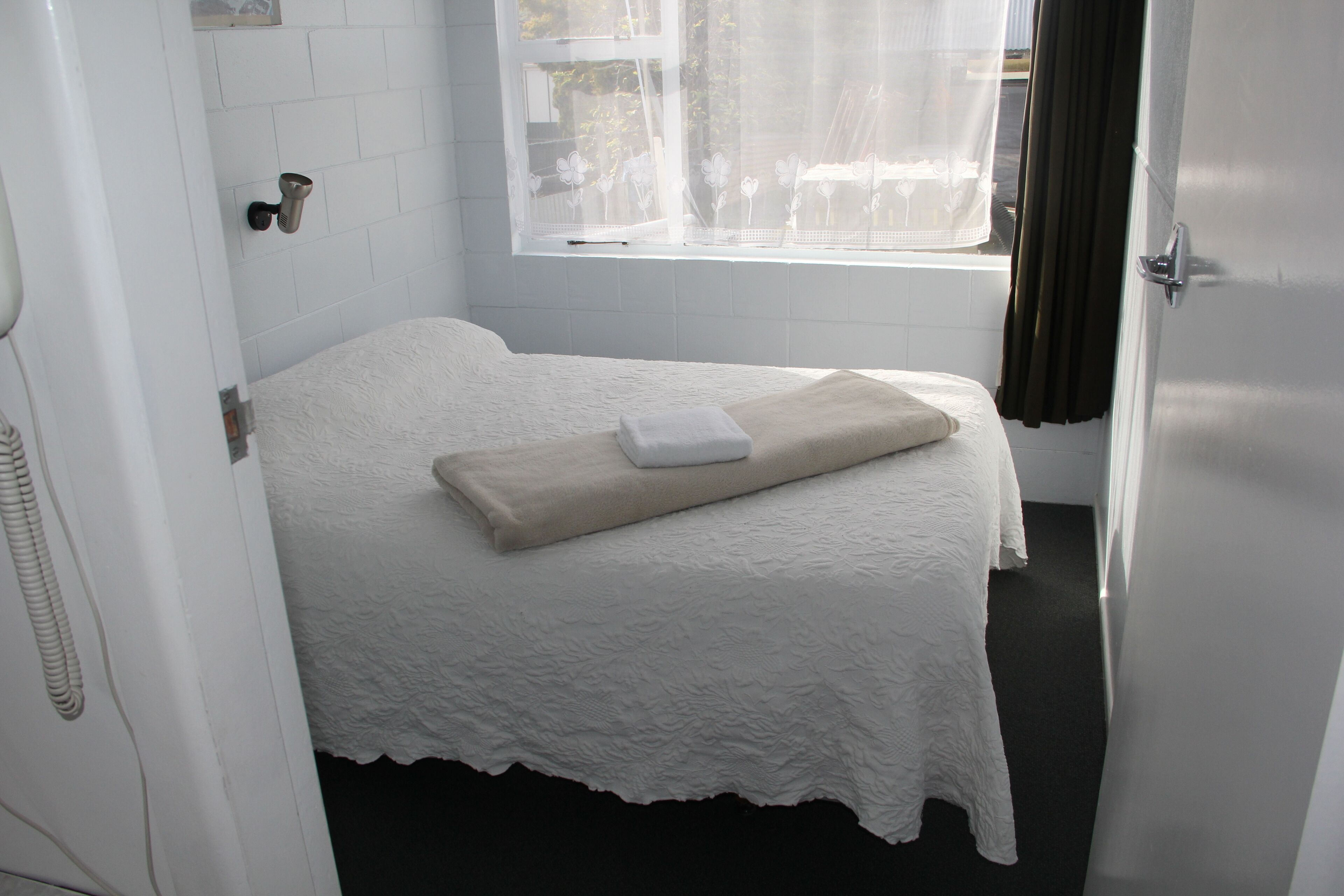 A small, neatly made bed with a white bedspread and a folded towel, a window with sheer curtains, and a white wall with a door.