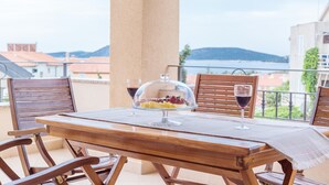A wooden table with a glass dome dessert stand, two wine glasses, and a view of a body of water and buildings in the background. - Luxury Apartments Iva (Vodice)