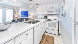 Una cocina moderna con armarios blancos, una isla grande con un lavabo, electrodomésticos de acero inoxidable, iluminación colgante y vistas al exterior.