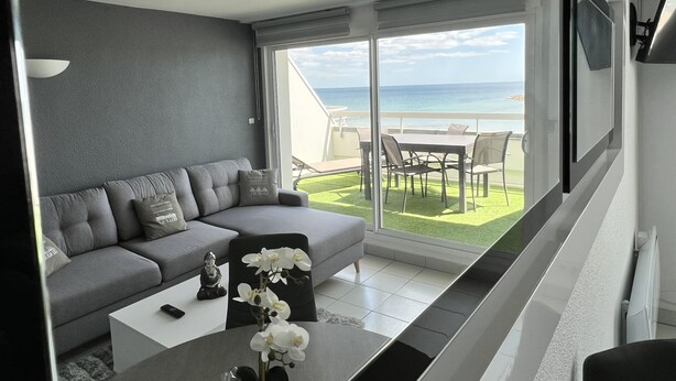 Un salón moderno con un sofá gris, una mesa de centro blanca y vista al océano a través de amplios ventanales correderas.