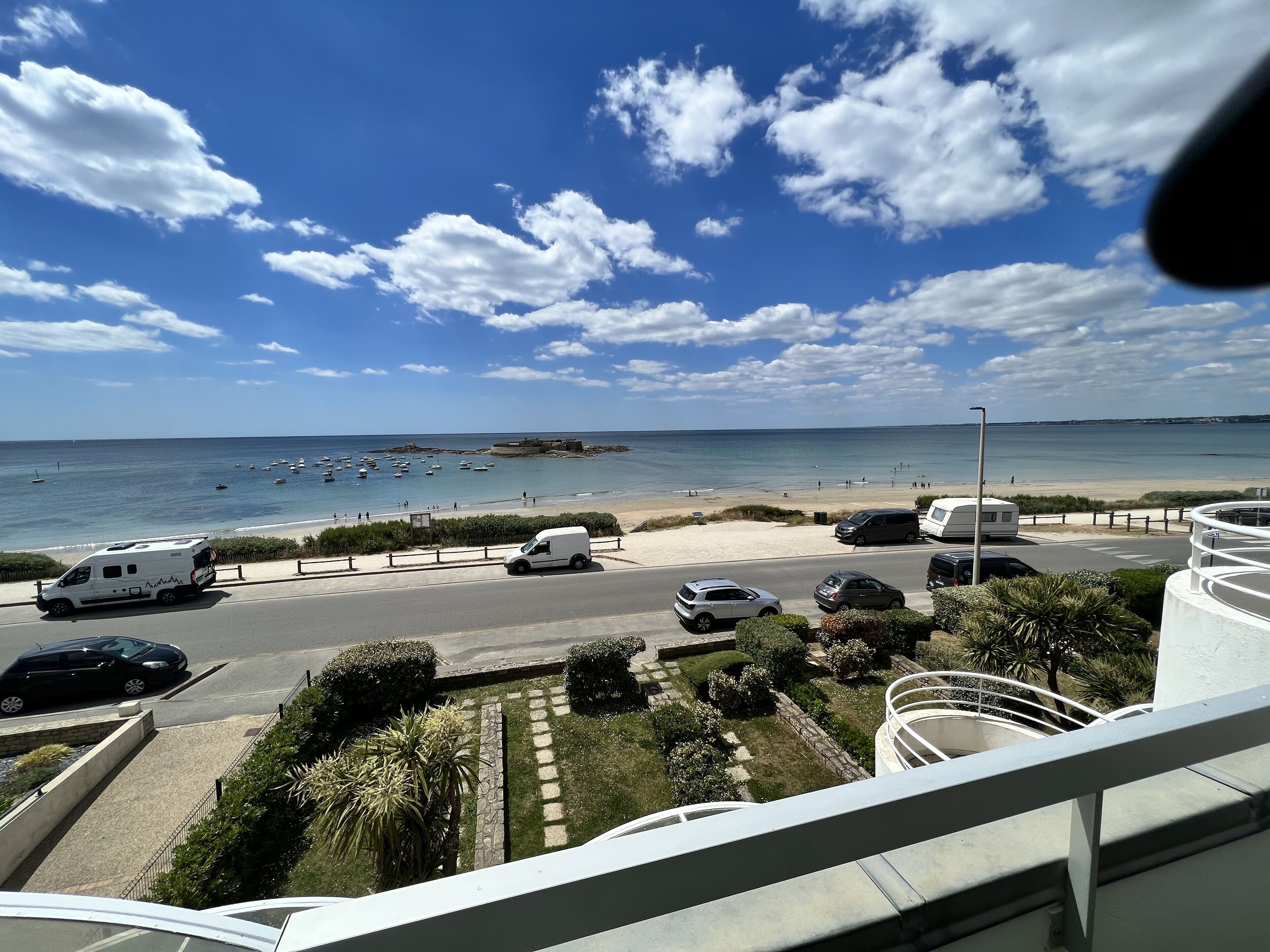 A coastal road with parked vehicles, a beach in the background, and a clear sky.