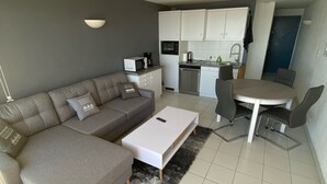 Una sala de estar moderna con un sofá gris, una mesa de centro blanca y un comedor con una mesa redonda y sillas. Se ve una cocina con gabinetes y electrodomésticos blancos al fondo.