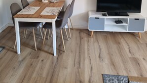 Uma sala de estar moderna com uma mesa de jantar de madeira, cadeiras cinzas, uma TV sobre um suporte branco e um lustre no teto.