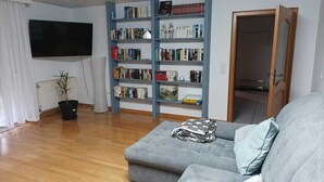 A living room with a grey sofa, a bookshelf, a radiator, a potted plant, and a television. - Large family-friendly apartment for 2 - 5 people on the first floor. (Rheinland-Pfalz - Beuren (Eifel))