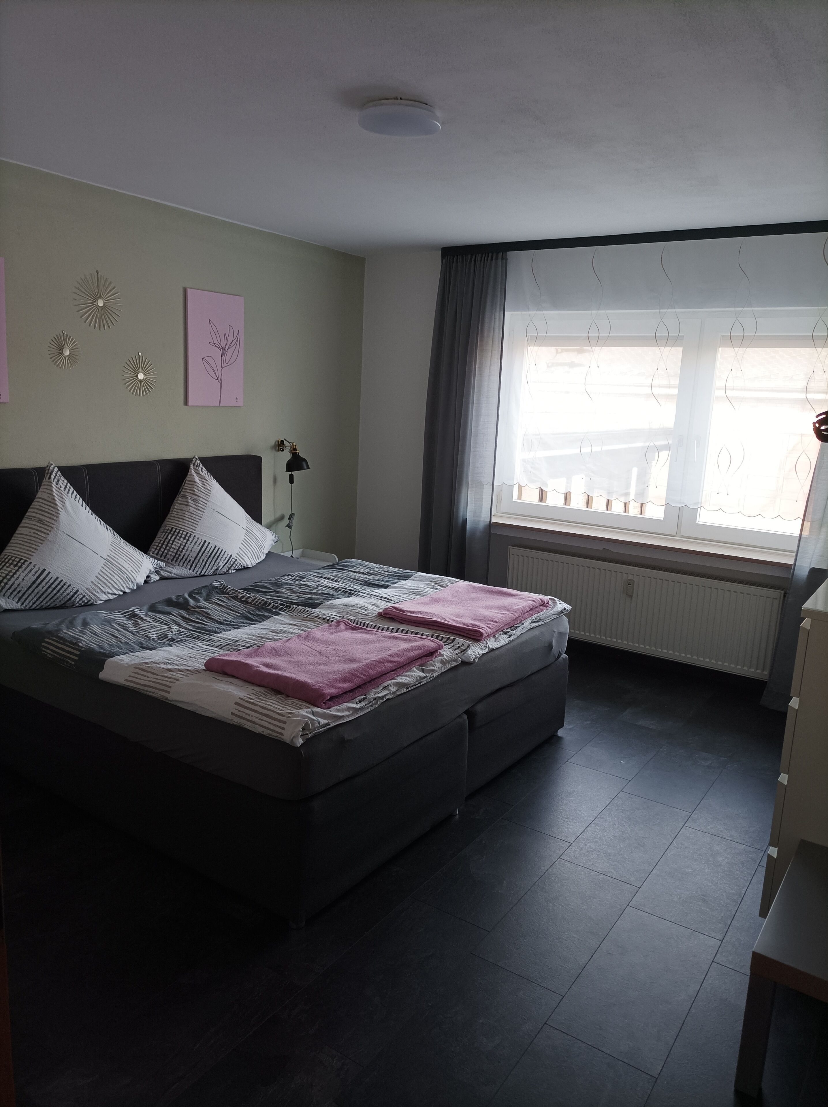 A bedroom with a bed, a bedside lamp, a window with curtains, and a radiator.