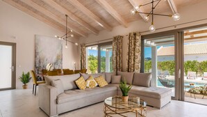 A spacious living room with a large sofa, a glass coffee table, and a view of a pool and outdoor seating area. - Villa Anemona - Zante Prime Heated Pool Villas  (Zakynthos)