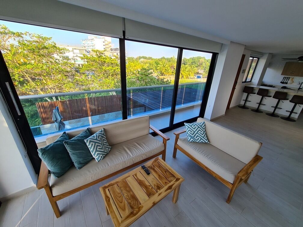 A modern living room with a large window, two sofas, a wooden coffee table, and a kitchen area with bar stools.