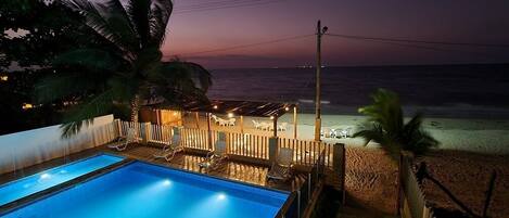A swimming pool with lounge chairs and umbrellas, a beach with a wooden fence, and a sunset over the ocean.