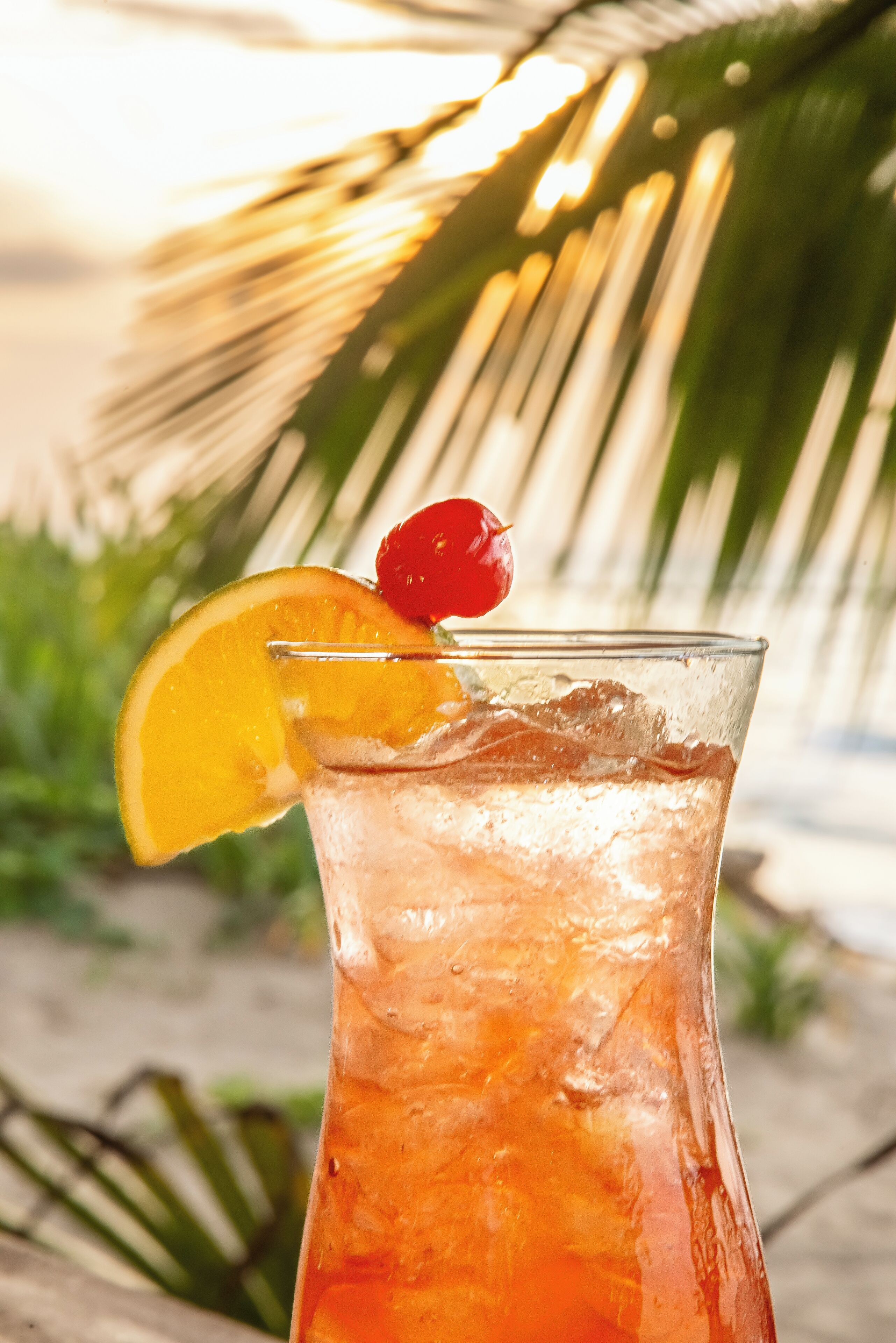 A glass of iced beverage with a cherry and orange slice garnish, set against a beach backdrop with palm trees.