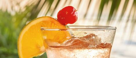 A glass of iced beverage with a cherry and orange slice garnish, set against a beach backdrop with palm trees.