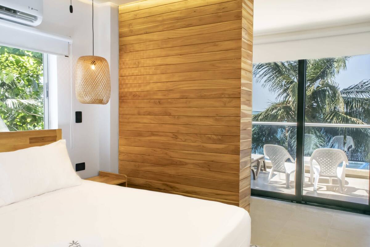 Une chambre moderne avec un mur d’accent en bois, un grand lit, un luminaire suspendu et une vue sur un balcon aménagé avec du mobilier d’extérieur.