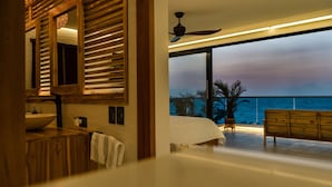 Un dormitorio moderno con un ventanal grande con vistas al océano, un ventilador de techo y un tocador de madera.