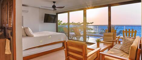 A room with a bed, a TV, a balcony with a view of the sea, and a dining area with chairs and a table.