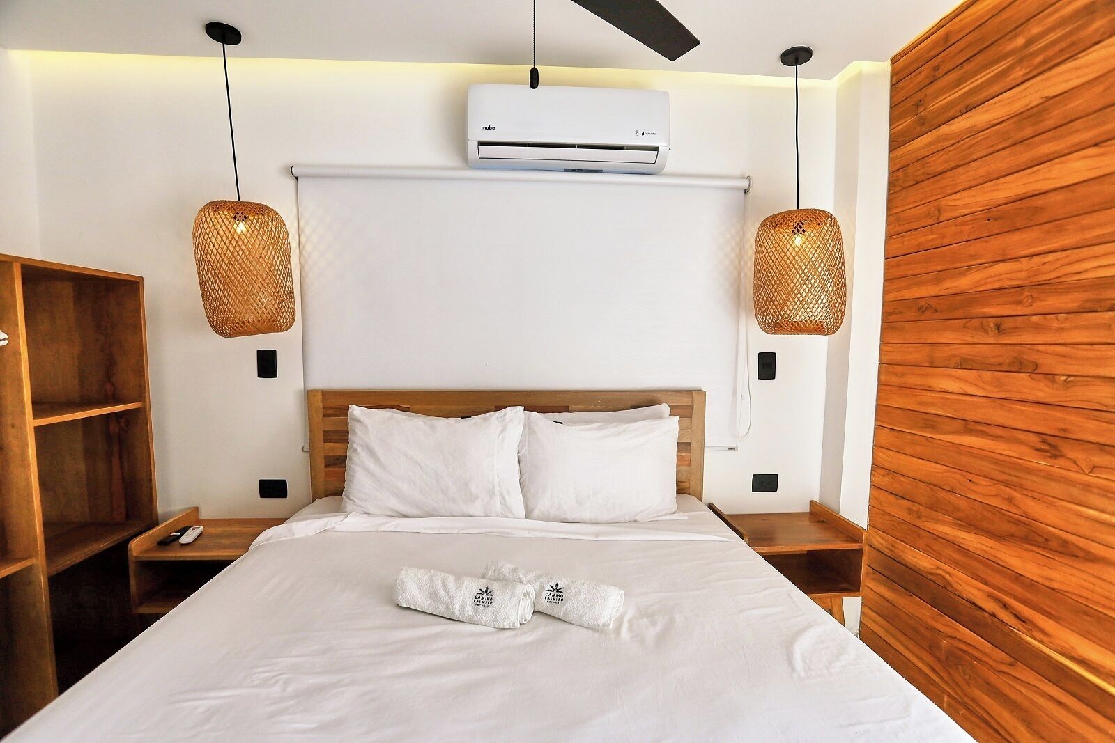 A hotel room with a wooden headboard, white bedding, a rolled towel, and two wicker lamps.