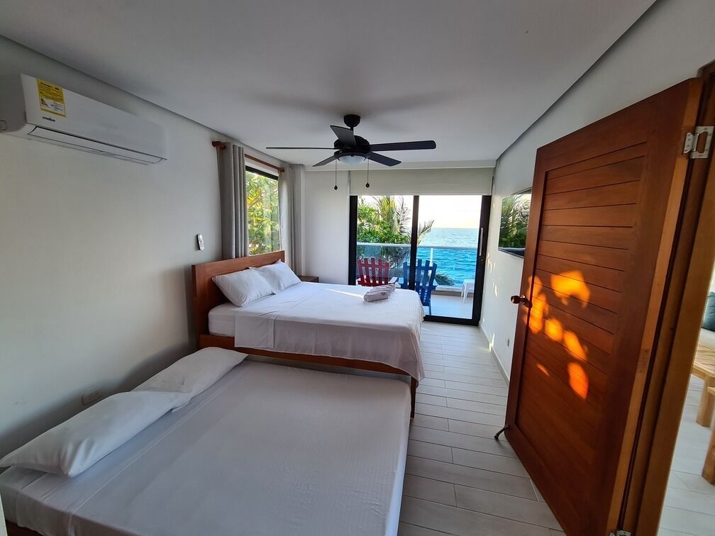 A room with two beds, a ceiling fan, and a view of the sea through a sliding door.