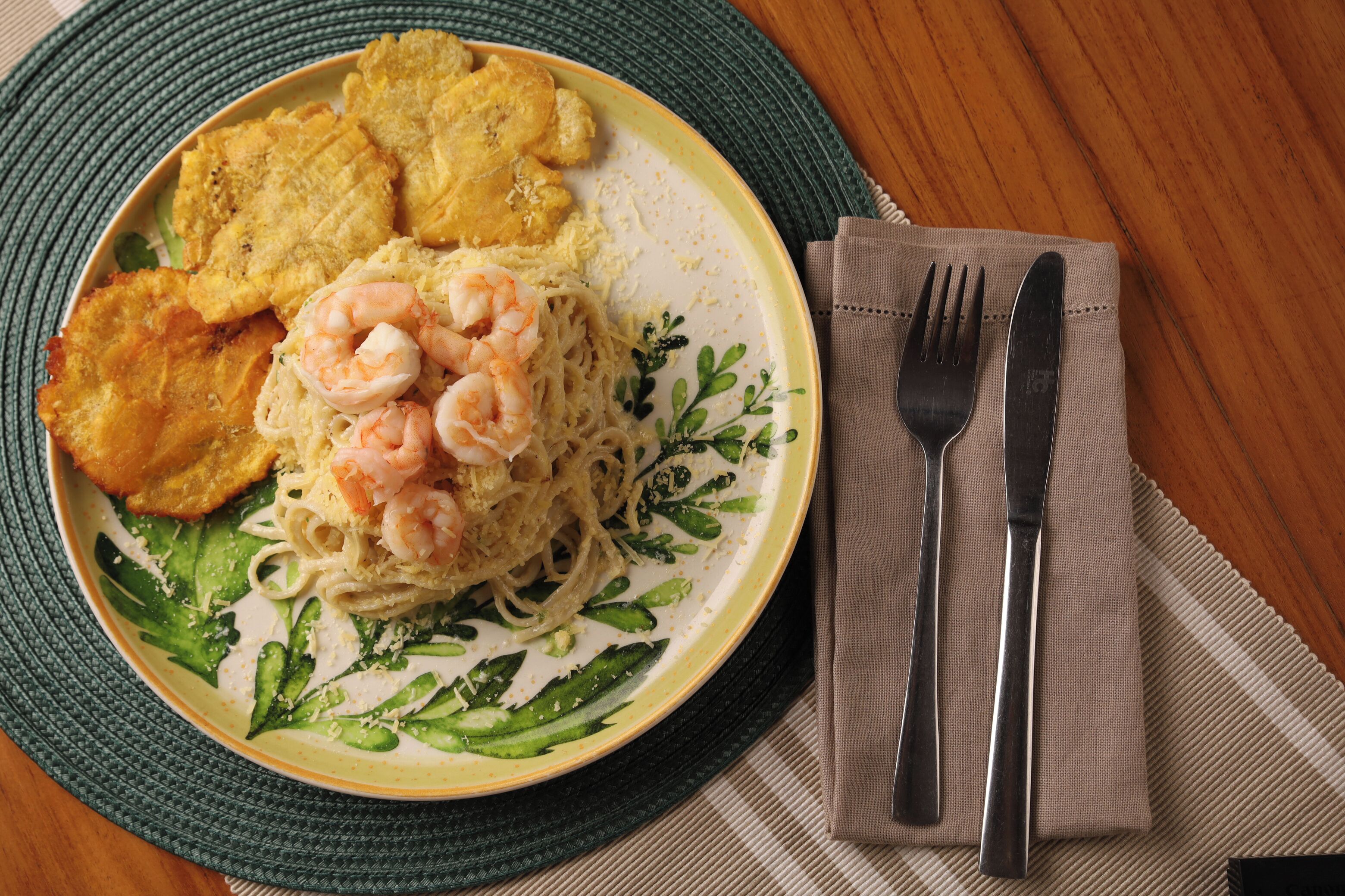 A plate of food with shrimp and noodles, accompanied by fried plantains, on a table with a placemat and cutlery.