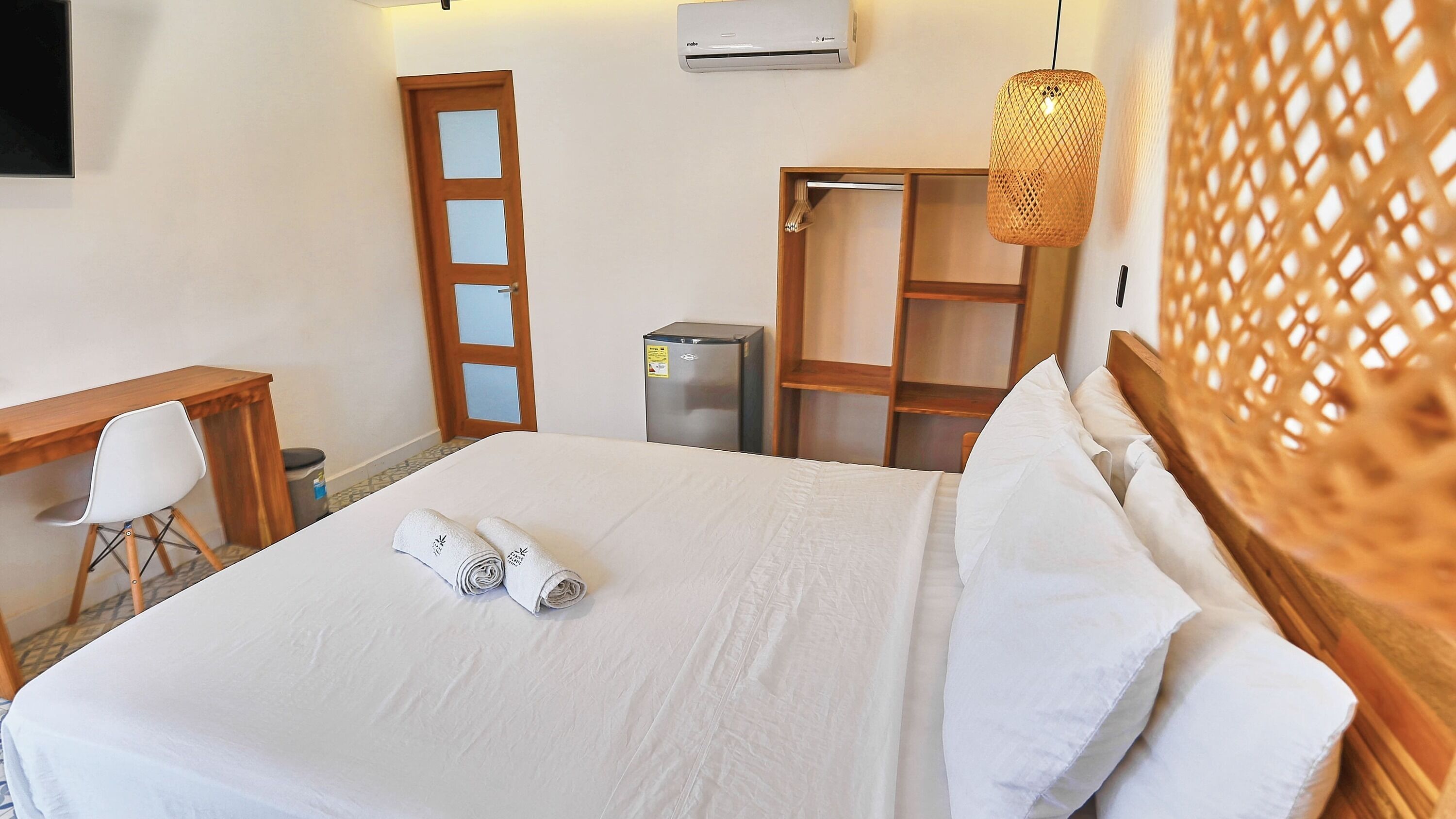 A neatly made bed with white linens, a wooden desk with a chair, a refrigerator, and a wardrobe.