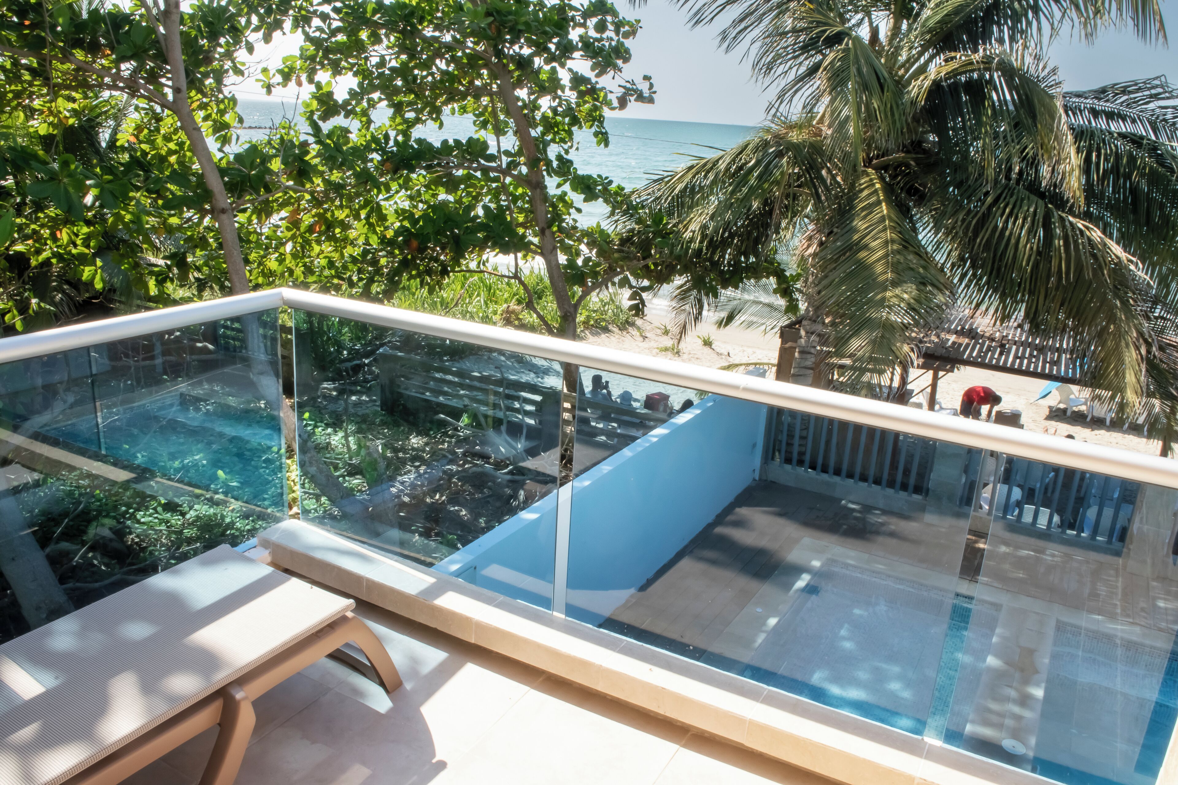 A glass-enclosed balcony with a pool, overlooking a beach and palm trees.