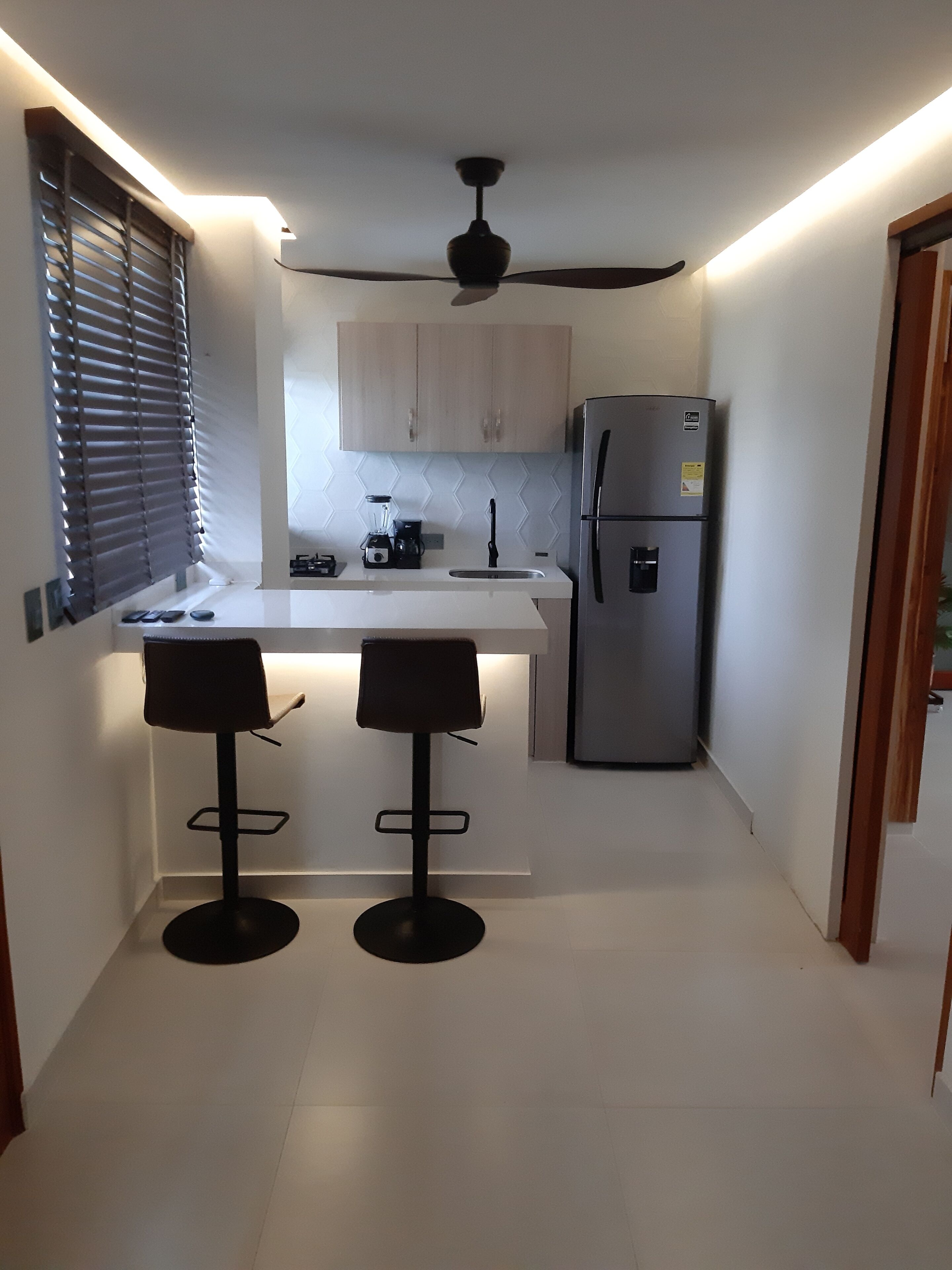 Una cocina moderna con una isla central, dos taburetes de bar, un refrigerador y un ventilador de techo.