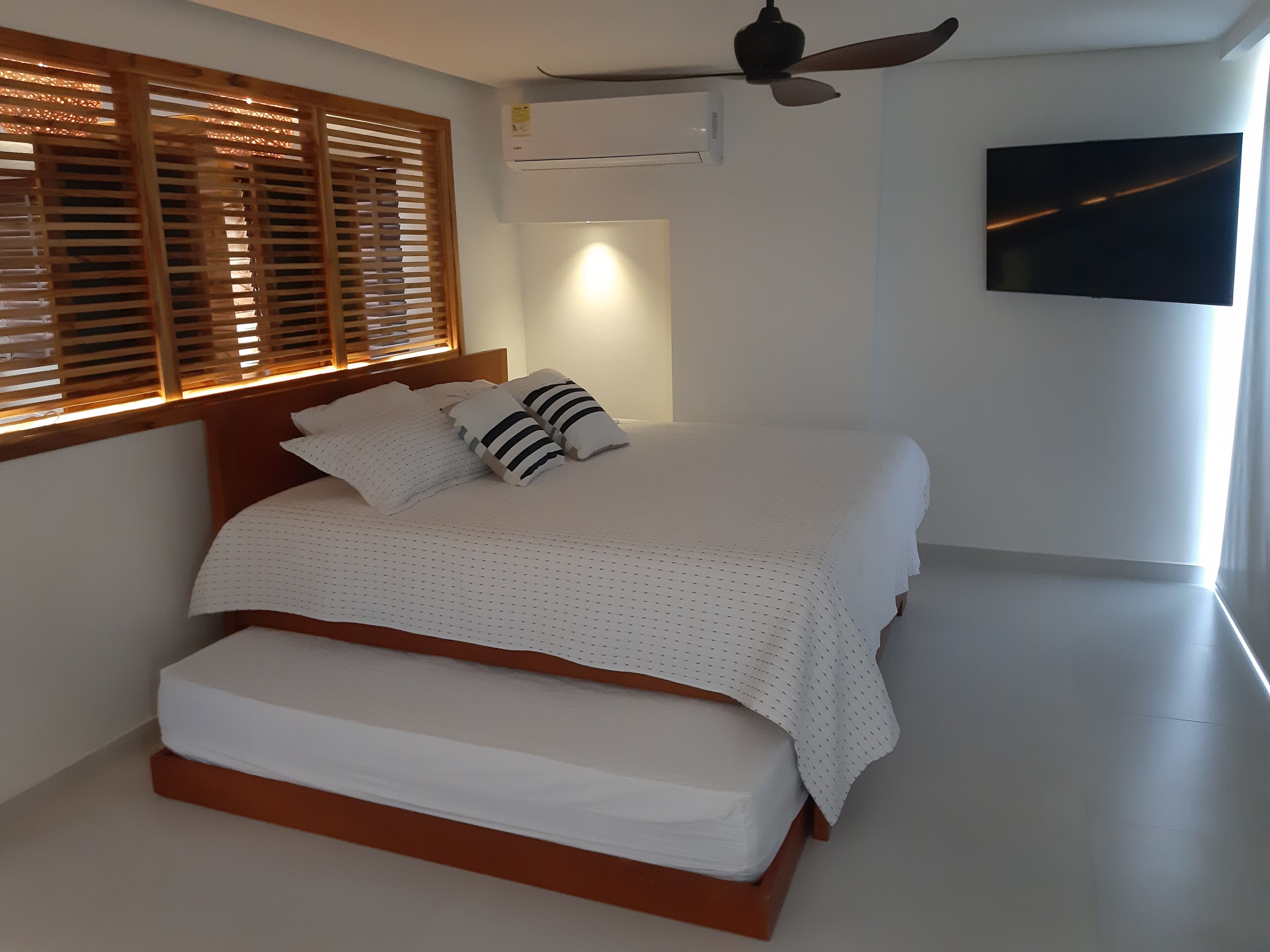 A bedroom with a bed, wooden headboard, white bedding, a ceiling fan, an air conditioning unit, and a TV on the wall.