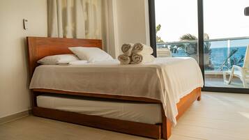 Habitación de hotel con cama de madera, ropa de cama blanca, vista al océano y un balcón con una silla.
