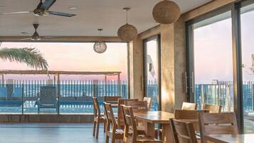 A spacious balcony with wooden furniture, large windows, and a view of the sea at sunset.