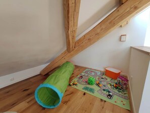 A room with a wooden floor, a green and blue tunnel, a play mat with vehicles, and a small orange container.