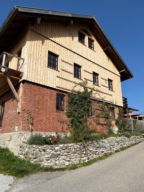 A two-story building with a wooden facade and a brick base, featuring a stone wall and a small garden area.