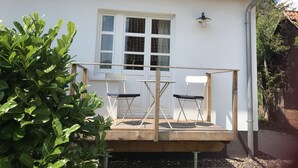 Uma casa branca com telhado de cerâmica, um deck de madeira, duas cadeiras e uma janela com cortinas.