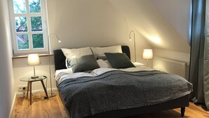 Quarto com cama, duas luminárias de cabeceira, janela e uma mesinha.