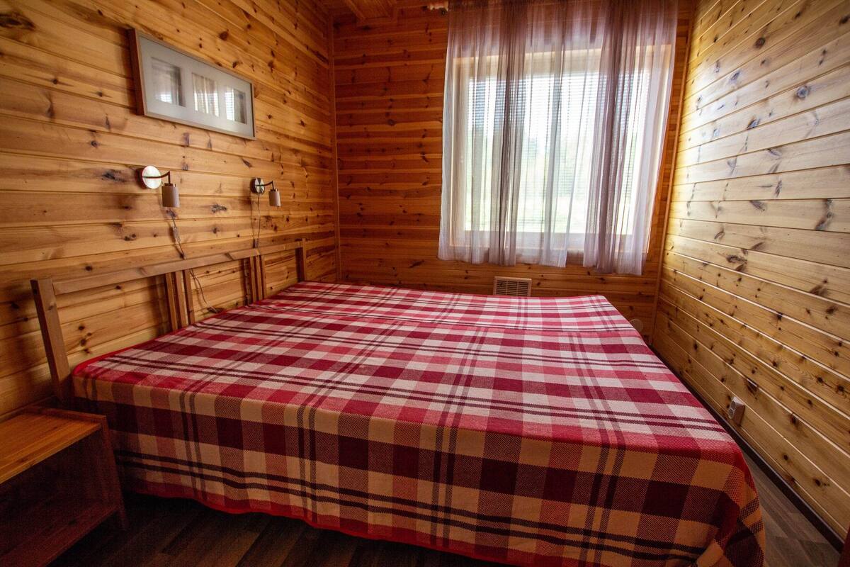 A wooden cabin room with a plaid bedspread, a framed picture on the wall, and a window with sheer curtains.