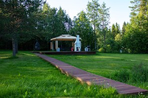 A wooden deck leading to a gazebo with a chimney, surrounded by lush greenery.