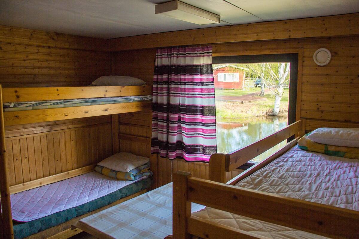 A cabin with three bunk beds, a window with a view of a pond and a red building, and a striped curtain.